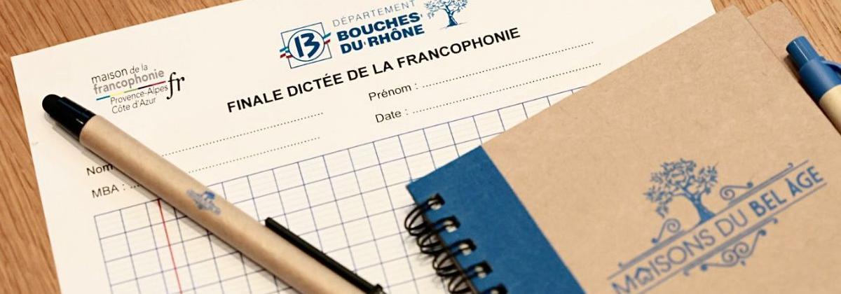Sur une table en bois reposent une feuille de dictée officielle du Département, un cahier bleu et kraft, ainsi qu’un stylo noir et beige. Le document comporte des espaces pour nom, prénom et date. L’ensemble évoque une préparation à un concours d’écriture et dans le cas présent, une dictée.