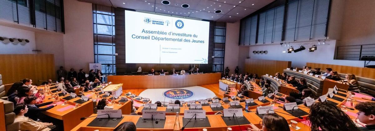 Salle de réunion officielle vue de l’arrière. De nombreux jeunes sont assis autour de pupitres en bois, regardant un grand écran affichant une assemblée d’investiture. Micros, dossiers et bouteilles d’eau visibles.