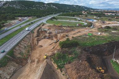 Vue aérienne d’un vaste chantier près d’une autoroute. Aucune personne visible. Plusieurs engins de chantier (pelles, camions, bulldozers) creusent et déplacent la terre. La scène est entourée de collines boisées et de quartiers résidentiels au loin. Le trafic roule sur plusieurs voies adjacentes.