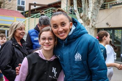 Une joueuse de l'Olympique de Marseille, souriante, pose aux côtés d'une jeune fille portant des lunettes et une veste noire, dans la cour d'un collège animé - Agrandir l'image 10 sur 16, fenêtre modale