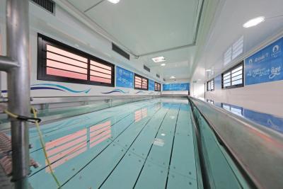 Piscine intérieure étroite avec eau claire, murs blancs, fenêtres à cadres noirs et banderoles informatives visibles. Il s'agit de l'intérieur d'un camion piscine. - Agrandir l'image 7 sur 8, fenêtre modale