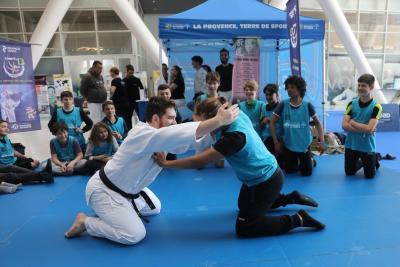 des ados assis sur un tatami en face d'un stand. un adolescent et un adulte se font face à genou, l'un en kimono ceinture noire, l'autre avec un dossard. Ils sont en pleine lutte. - Agrandir l'image 4 sur 9, fenêtre modale