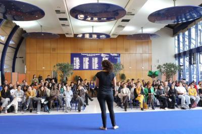 Grande salle lumineuse avec sol bleu et plafond décoré de disques bleus. Une personne debout face à un large public assis sur des chaises fait un discours. Derrière, mur en bois avec panneau bleu listant des noms. Deux grands arbres décoratifs sont visibles. Les participants regardent la personne qui semble présenter ou animer. - Agrandir l'image 2 sur 5, fenêtre modale