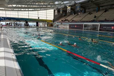 Grand bassin intérieur avec lignes de nage colorées. Plusieurs nageurs équipés de bonnets et lunettes nagent dans des couloirs. Gradins visibles en arrière-plan avec quelques spectateurs. Structure moderne avec toit métallique et larges baies vitrées. - Agrandir l'image 5 sur 5, fenêtre modale