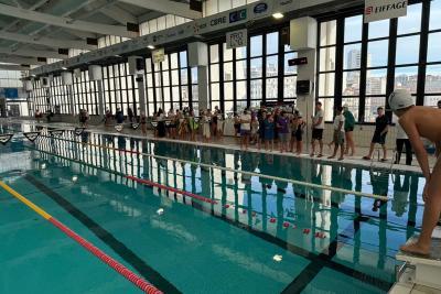 Piscine intérieure avec couloirs de nage. Une personne en bonnet se tient prête à plonger sur un plot au premier plan. En arrière, groupe aligné le long du bassin, semblant attendre leur tour. Grandes fenêtres laissant entrer la lumière, ambiance de compétition. - Agrandir l'image 4 sur 5, fenêtre modale