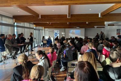 Même salle, vue élargie : rangées de chaises occupées par de nombreuses personnes, écran de projection au fond affichant une image, table avec intervenants à gauche. Lumière naturelle abondante, sol en bois, ambiance de conférence ou présentation. - Agrandir l'image 2 sur 5, fenêtre modale