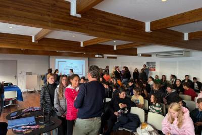 Salle avec public assis et quelques personnes debout au centre, semblant échanger. À l’arrière, écran projetant « PRIX ALFRED NAKACHE 2025 ». Sur une table proche, médailles disposées. Atmosphère de remise de prix ou cérémonie. - Agrandir l'image 3 sur 5, fenêtre modale