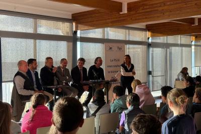 Salle lumineuse avec grandes baies vitrées et poutres en bois. Six personnes assises derrière une table haute avec micros, une autre debout près d’un panneau indiquant « Le FSJU vous souhaite la bienvenue ». Devant, public assis sur des chaises, tourné vers les intervenants. - Agrandir l'image 1 sur 5, fenêtre modale