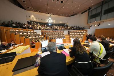 Vue depuis la tribune de l'hémicycle. À l’avant, des personnes assises derrière des pupitres avec micros et documents. Certaines portent l’écharpe jaune et bleue. En face, des gradins remplis de jeunes et adultes. L’écran central et les panneaux en bois renforcent le cadre institutionnel. - Agrandir l'image 3 sur 5, fenêtre modale