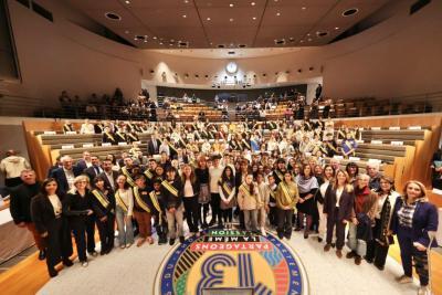 Dans la salle des séances publiques, l'hémicycle est rempli de nombreuses personnes, principalement des jeunes, debout et regroupés pour une photo collective. Beaucoup portent une écharpe jaune et bleue. Les gradins en bois et sièges gris sont visibles, ainsi qu’un sol décoré avec un grand logo circulaire « 13 ». L’ambiance est solennelle et institutionnelle. - Agrandir l'image 5 sur 5, fenêtre modale
