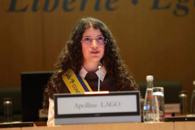 La nouvelle Présidente du Conseil Départemental des Jeunes est assise derrière un pupitre avec une plaque nominative « Apolline LAGO ». Devant elle, un micro et deux bouteilles d’eau. En arrière-plan, un mur en bois avec les mots « Liberté • Égalité ». L’écharpe jaune et bleue « Conseil Départemental des Jeunes » est portée en bandoulière. - Agrandir l'image 2 sur 5, fenêtre modale
