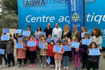 Un groupe d’enfants tient des diplômes bleus devant un grand panneau « Centre aquatique » accroché au camion-piscine et une bannière « Département Bouches-du-Rhône ». Ils sont alignés en extérieur sur sol gris, entourés d’adultes. - Agrandir l'image 3 sur 5, fenêtre modale