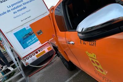 Un véhicule orange avec logo « Peypin » est garé, porte ouverte, à côté du camion-piscine affichant « La première école de natation itinérante au monde ». Un tuyau relie les deux. Sol en bitume, arrière-plan avec structure métallique et affiche bleue sur la natation. - Agrandir l'image 5 sur 5, fenêtre modale