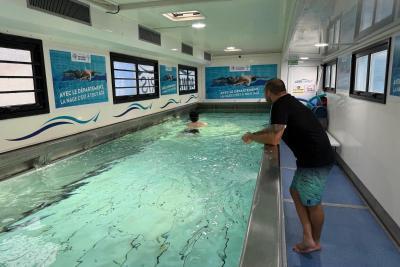 Intérieur du camion-piscine. Un maître nageur en short se tient au bord d’un bassin intérieur, observant un enfant qui nage. L’eau est turquoise, murs blancs ornés d’affiches sur la natation. Fenêtres latérales, sol bleu antidérapant, ambiance pédagogique. - Agrandir l'image 2 sur 5, fenêtre modale