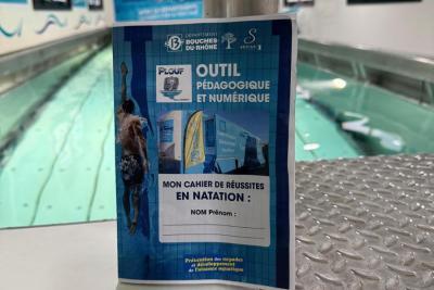 À l’intérieur d’un espace lumineux qui est l'intérieur du camion-piscine, un bassin rectangulaire rempli d’eau claire. Au premier plan, un livret bleu « Outil pédagogique et numérique – Mon cahier de réussites en natation » posé sur une surface métallique. Affiches éducatives sur les murs. - Agrandir l'image 1 sur 5, fenêtre modale