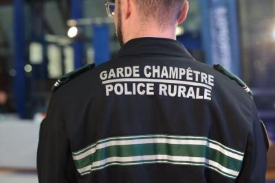Un adulte de dos porte une veste noire avec bandes vertes et blanches, marquée “GARDE CHAMPÊTRE POLICE RURALE”. L’environnement est un espace intérieur éclairé, probablement un hall. D’autres personnes en uniforme sont visibles en arrière‑plan. La scène se déroule pendant la cérémonie des voeux cérémonie des voeux au personnel des forces de l'ordre et de secours des Bouches-du-Rhône. - Agrandir l'image 2 sur 18, fenêtre modale