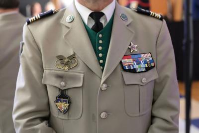 Un adulte porte un uniforme militaire beige avec une veste ornée de plusieurs insignes : un aigle doré ailé, un écusson noir et or, ainsi qu’un ensemble de décorations colorées sur la poitrine. Il porte une chemise blanche avec cravate noire et un gilet vert. La scène se déroule en intérieur, avec d'autres personnes en tenue officielle visibles en arrière‑plan. Il s'agit de la cérémonie des voeux au personnel des forces de l'ordre et de secours des Bouches-du-Rhône. - Agrandir l'image 13 sur 18, fenêtre modale