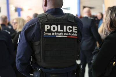 Un adulte en tenue sombre est vu de dos, portant un gilet tactique noir marqué “POLICE Groupe de Sécurité et de Proximité” avec un liseré tricolore. Il se trouve dans un hall fréquenté par plusieurs personnes en uniforme, en pleine discussion. - Agrandir l'image 11 sur 18, fenêtre modale