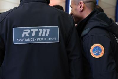 Un adulte vu de dos porte une veste sombre avec un large marquage “RTM – ASSISTANCE • PROTECTION” en blanc sur un panneau gris. À côté, un autre adulte en uniforme foncé présente un écusson rond orange et bleu sur la manche. Les deux se tiennent dans un espace intérieur fréquenté par d’autres personnes en tenue professionnelle, évoquant un rassemblement ou une coordination de services. Il s'agit de la cérémonie des voeux au personnel des forces de l'ordre et de secours. - Agrandir l'image 14 sur 18, fenêtre modale