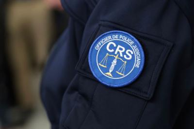 Un adulte en uniforme sombre est montré de près, centré sur la manche. Un écusson rond bleu et blanc y est fixé, portant la mention “OFFICIER DE POLICE JUDICIAIRE – CRS”. L’emblème représente une balance et un flambeau dorés. L’environnement semble être un espace intérieur avec d’autres personnes en tenue professionnelle en arrière‑plan, dans un contexte officiel. Il s'agit de la cérémonie des vœux au personnel des forces de l'ordre et de secours. - Agrandir l'image 15 sur 18, fenêtre modale