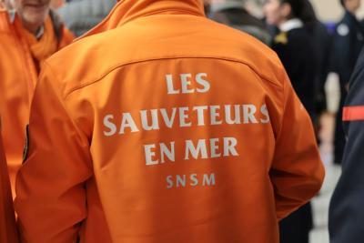 Un adulte vu de dos porte une veste orange vif avec l’inscription “LES SAUVETEURS EN MER SNSM”. La scène se situe dans un espace intérieur animé où d’autres personnes en uniforme semblent échanger. L’atmosphère évoque un regroupement professionnel. Il s'agit de la cérémonie des voeux au personnel des forces de l'ordre et de secours des Bouches-du-Rhône. - Agrandir l'image 12 sur 18, fenêtre modale