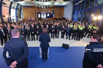 Un large rassemblement se tient dans une salle moderne et lumineuse. Au centre, une personne en tenue professionnelle parle derrière un pupitre face à une foule composée majoritairement d’adultes en uniformes (police, gendarmerie, sécurité, services municipaux). Les participants sont alignés, écoutant attentivement. L’espace est vaste, avec de grandes baies vitrées et un éclairage bleu. - Agrandir l'image 18 sur 18, fenêtre modale