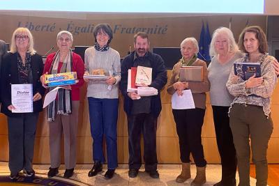 Un groupe d’adultes se tient debout devant un mur portant la devise républicaine. Chacun tient un lot : jeux, livres ou boîtes. Ils sont alignés côte à côte comme lors d’une remise de prix, dans une salle officielle au décor institutionnel. - Agrandir l'image 5 sur 5, fenêtre modale
