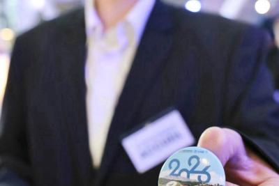 Personne tenant un badge circulaire « 2026 » en avant‑plan dans un espace événementiel éclairé - Agrandir l'image 1 sur 20, fenêtre modale