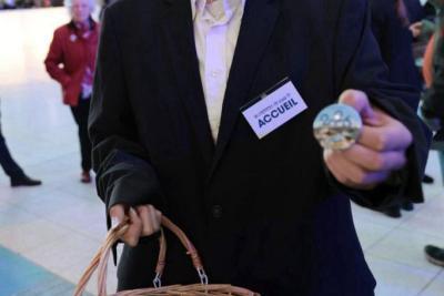 Personne du service accueil présentant un badge « 2023 » et tenant un panier rempli de badges dans un hall événementiel - Agrandir l'image 2 sur 20, fenêtre modale