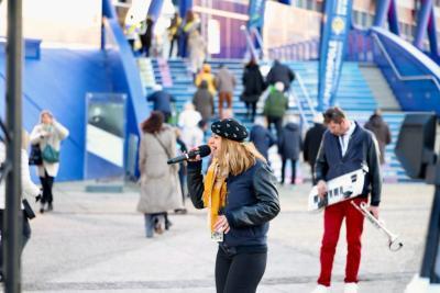Chanteuse interprétant un titre au micro en extérieur devant un bâtiment bleu, accompagnée d’un musicien jouant d’un keytar parmi les passants - Agrandir l'image 4 sur 20, fenêtre modale