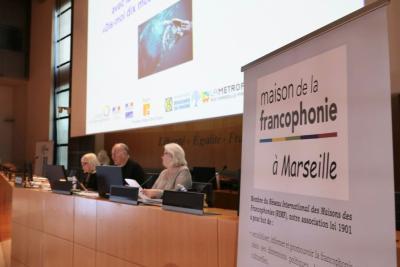 Dans un amphithéâtre institutionnel, trois personnes sont assises derrière une table équipée de micros et d’ordinateurs. Un grand écran projette une présentation. À droite, un panneau annonce « maison de la francophonie à Marseille ». L’ambiance est formelle et organisée. - Agrandir l'image 1 sur 5, fenêtre modale