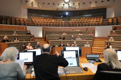 Une grande salle de conférence en gradins accueille des adultes assis, dispersés sur plusieurs rangées. Devant eux, trois personnes sont installées à une table équipée d’ordinateurs et de micros. L’ambiance est studieuse, éclairée par de larges spots, et un écran est visible au mur du fond. - Agrandir l'image 2 sur 5, fenêtre modale