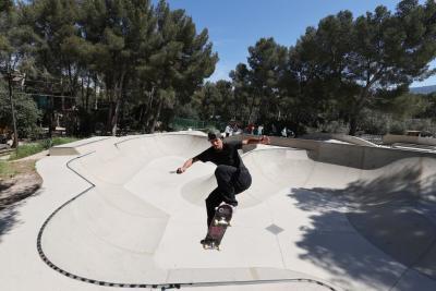 Un adulte pratique le skateboard dans une grande cuvette en béton d’un skatepark. La personne est en mouvement, légèrement accroupie sur la planche, les bras écartés pour l’équilibre. Plusieurs autres personnes sont visibles en arrière-plan, assises ou debout autour du skatepark. L’environnement est extérieur, entouré de nombreux arbres, avec une forte luminosité indiquant une journée ensoleillée. - Agrandir l'image 3 sur 3, fenêtre modale