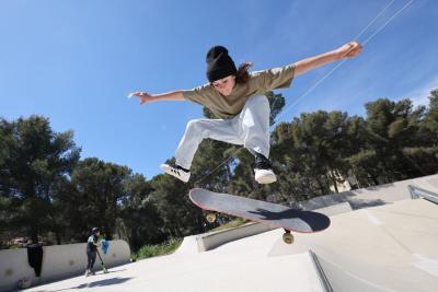 Une adolescente est photographiée en plein saut au-dessus d’une rampe de skate. Le skateboard est décollé du sol sous les pieds, le corps penché en avant avec les bras écartés pour l’équilibre. La scène se déroule dans un skatepark extérieur entouré d’arbres. En arrière-plan, un autre enfant se déplace avec une trottinette. Le ciel est bleu et dégagé, renforçant l’impression de mouvement. - Agrandir l'image 2 sur 3, fenêtre modale