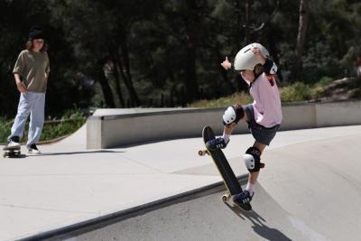 Une jeune enfant, équipée d’un casque et de protections aux genoux et coudes, fait du skateboard dans un skatepark en béton. L’enfant roule sur le bord incliné d’une rampe, les bras levés pour garder l’équilibre. À l’arrière-plan, un adolescent se tient sur un skateboard et regarde la scène. Le skatepark est entouré d’arbres, sous une lumière vive de plein jour. - Agrandir l'image 1 sur 3, fenêtre modale