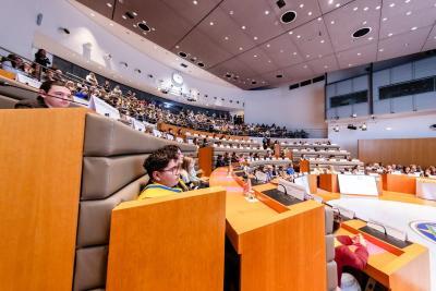 Grande salle hémicirculaire remplie d’adolescents et d’adultes assis sur plusieurs niveaux. Tous écoutent une séance officielle, entourés de pupitres en bois, micros, écrans et dossiers colorés. - Agrandir l'image 2 sur 6, fenêtre modale