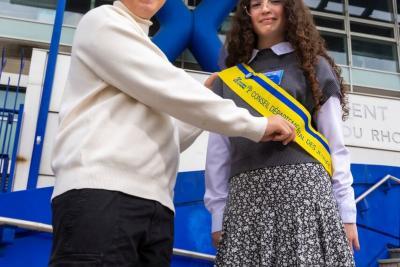 Deux adolescents debout devant un bâtiment moderne bleu. L’un tend une écharpe officielle jaune et bleue à l’autre. Ils sont sur des marches, près d’un drapeau et d’une façade administrative. - Agrandir l'image 4 sur 6, fenêtre modale