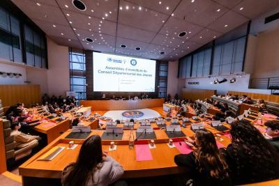 Salle de réunion officielle vue de l’arrière. De nombreux jeunes sont assis autour de pupitres en bois, regardant un grand écran affichant une assemblée d’investiture. Micros, dossiers et bouteilles d’eau visibles. - Agrandir l'image 1 sur 6, fenêtre modale