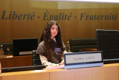 Jeune fille assise seule à un pupitre dans une salle officielle. Devant elle, un panneau portant un nom. Derrière, les mots “Liberté Égalité Fraternité” sur un mur boisé et des écrans de séance. - Agrandir l'image 5 sur 6, fenêtre modale