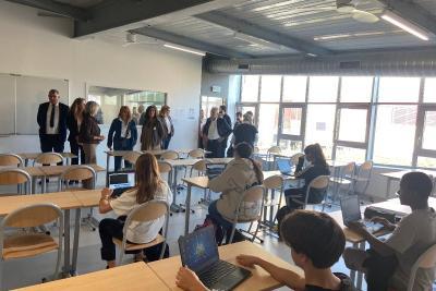 Des élèves sont assis à leur bureau dans une salle de classe, travaillant sur des ordinateurs portables. À l’arrière-plan, un groupe d’adultes observe la scène, probablement en visite pédagogique. - Agrandir l'image 2 sur 6, fenêtre modale