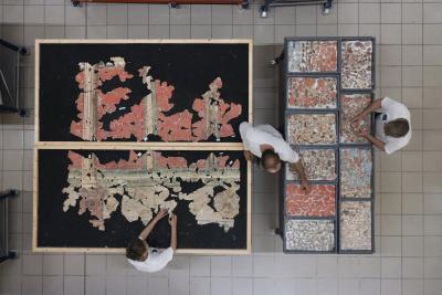 des personnes reconstituent une fresque, morceaux par morceaux sur une table de fond noir. A côté, les morceaux de la fresque ont été triés dans différents bacs, par couleurs. - Agrandir l'image 7 sur 14, fenêtre modale