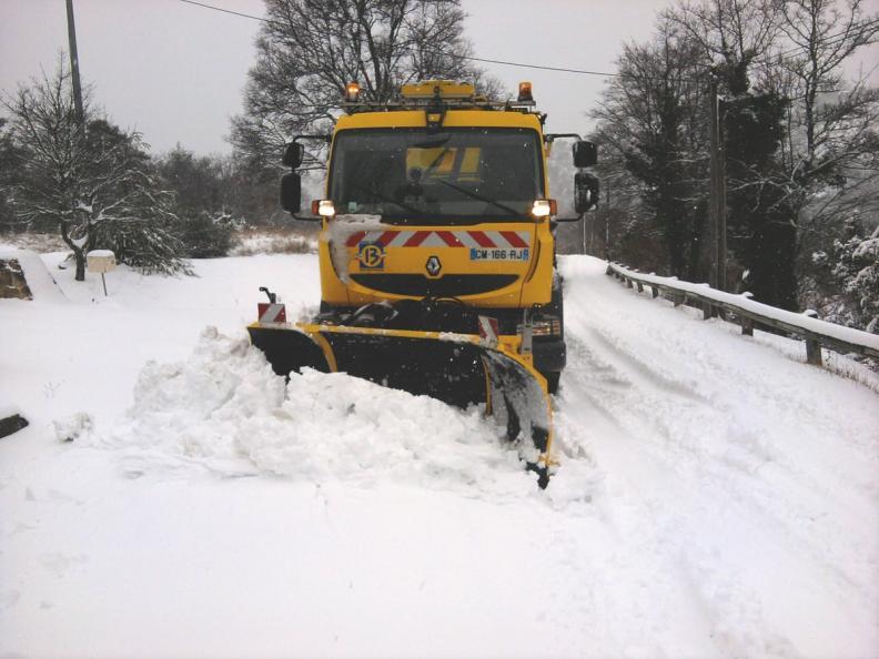 Sur une route enneigée bordée d’arbres nus, un camion chasse-neige jaune avance en poussant un large amas de neige. Aucun piéton visible. Le véhicule occupe le centre de l’image, entouré d’un paysage hivernal calme et recouvert d’une épaisse couche blanche.