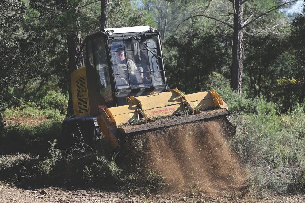 Engin forestier jaune et noir en action, projetant de la terre et des végétaux broyés sur un chemin dans une zone boisée.